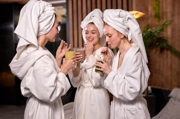 Three women in white bathrobes and towels on their heads enjoying drinks at a spa.